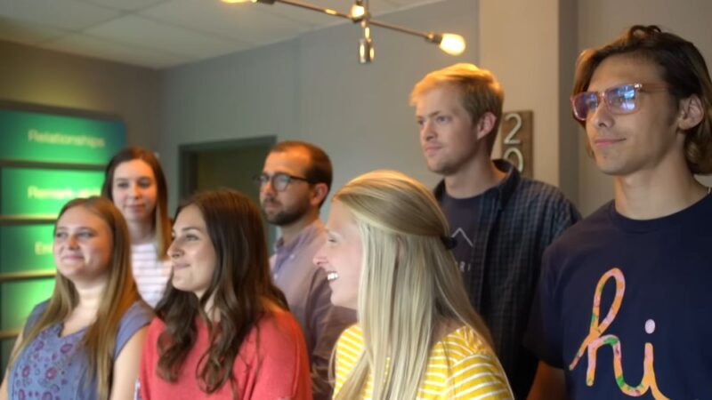 Grupa mladih zaposlenih stoji u kancelariji, nasmejani i okrenuti ka sagovorniku tokom internog izazova