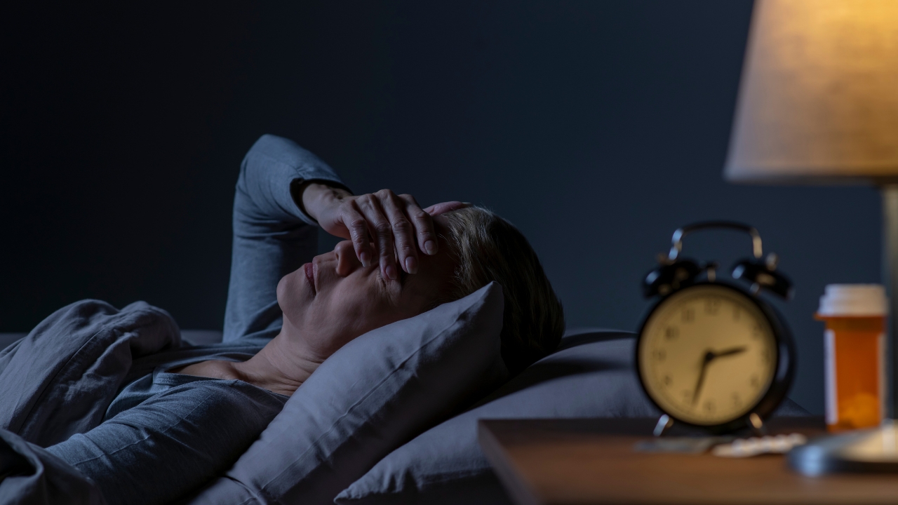 Žena leži budna u krevetu tokom noći pored uključenog sata i lampe