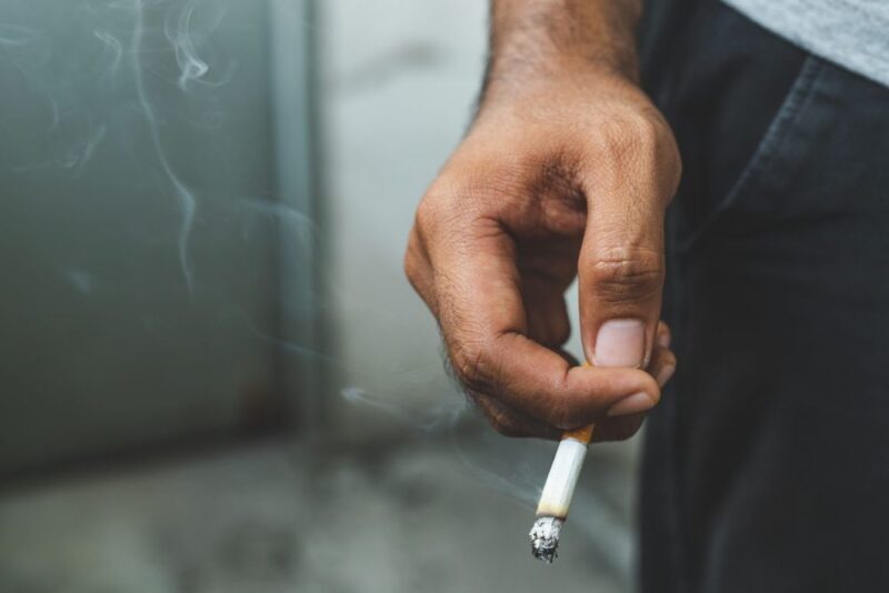 Krupan plan ruke muškarca koji drži zapaljenu cigaretu dok se dim širi oko nje.